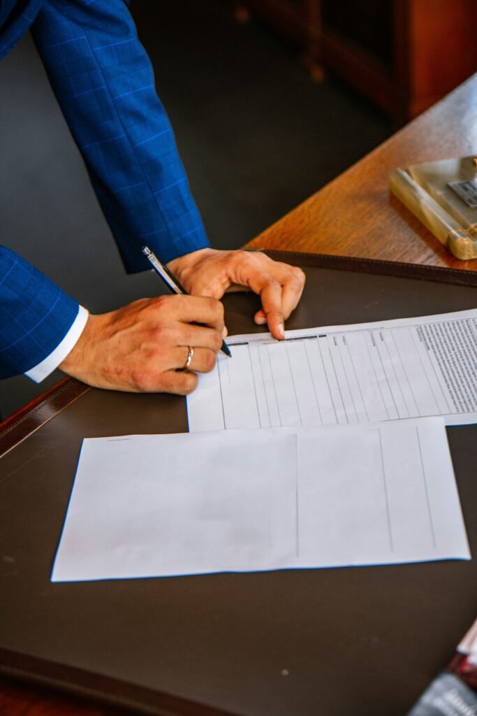 Une personne en costume bleu signe un document sur un bureau avec un stylo, avec des feuilles de papier vierges supplémentaires à proximité.