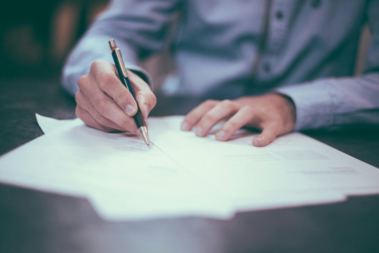 Une personne en chemise bleue écrit sur des documents papier avec un stylo à un bureau.
