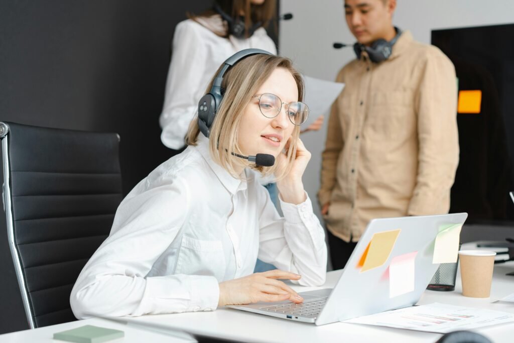 Une femme portant un casque travaille sur un ordinateur portable à un bureau avec des notes autocollantes, tandis que deux collègues avec des casques parlent en arrière-plan.